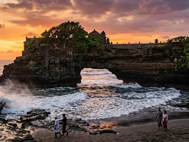 Pemandangan tebing karang ikonik Pura Batu Bolong di Tanah Lot, Bali, dengan lubang lengkungan alami di bawah tebing yang dialiri ombak laut berbusa putih. Di atas tebing karang tersebut berdiri bangunan pura yang dikelilingi pepohonan hijau. Latar belakang menampilkan langit senja dramatis dengan perpaduan warna jingga keemasan dan keunguan, sementara di tepi pantai berpasir hitam terlihat beberapa pengunjung sedang berjalan santai menikmati suasana sore.