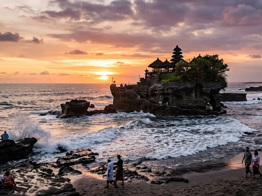 Pemandangan megah Pura Tanah Lot di Bali yang bertengger di atas bongkahan batu karang besar di tengah laut saat matahari terbenam. Langit dihiasi perpaduan warna jingga, emas, dan keunguan dengan awan yang dramatis. Di bagian bawah, ombak laut berbusa putih menghantam bibir pantai berpasir hitam, tempat beberapa pengunjung tampak berjalan santai menikmati suasana sore.